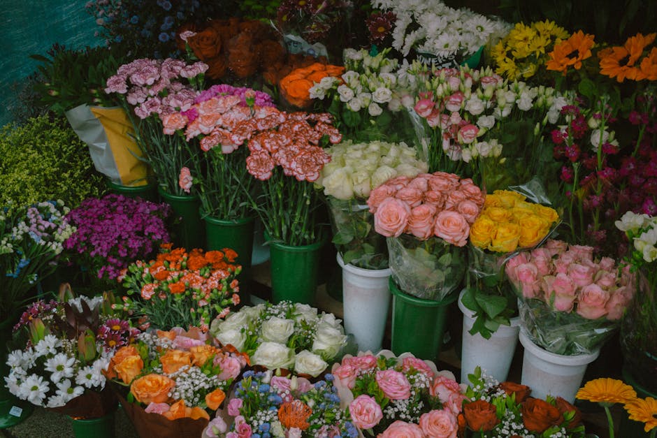 A vibrant display of various fresh flowers arranged in bouquets and vases. The arrangement includes pink roses, white roses, yellow roses, orange blossoms, and clusters of small daisy-like white flowe