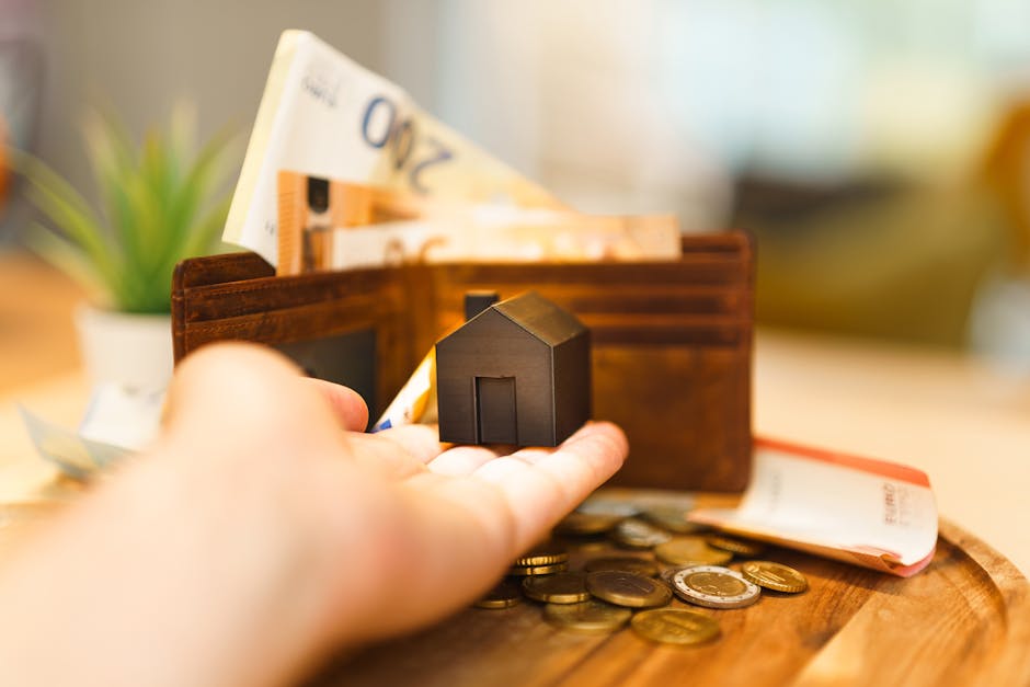 A close-up view of a person's hand holding a small black house-shaped object in front of a brown wooden wallet containing folded banknotes and scattered coins on a wooden surface. In the background, t