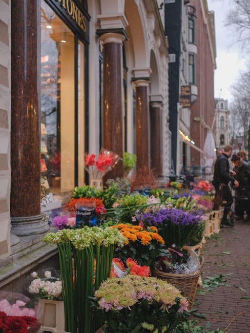 A vibrant flower stall outside a florist shop on Streatham High Road displays a diverse selection of fresh flowers arranged in wicker baskets and floral vases. The arrangement includes bright orange m