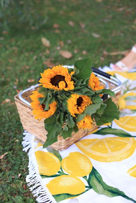 A bouquet of bright yellow sunflowers with dark centers and large green leaves, arranged in a rustic wicker basket. The basket rests on a floral-printed tablecloth with a lemon pattern, featuring yell